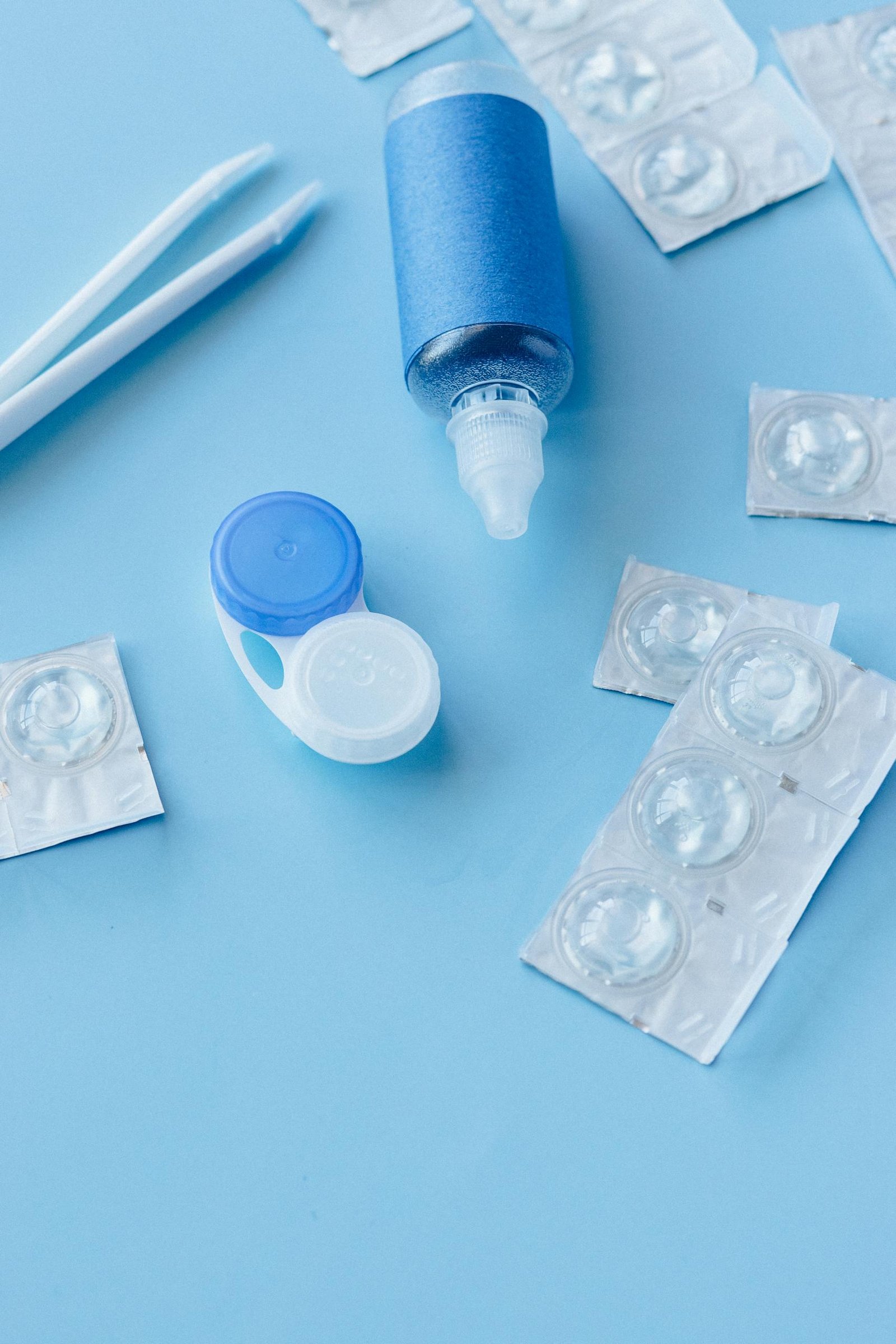 Flat lay image of a contact lens care kit including solution, lens container, and tweezers on a blue background.